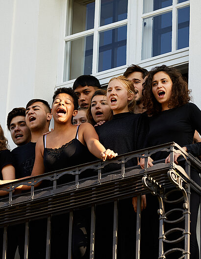 Eine Gruppe Jugendlicher singt auf einem Balkon.