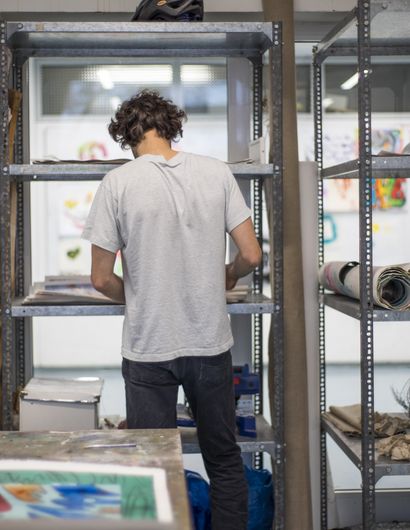 Ein Mann steht vor Regalen mit zusammengerollten Bildern. In der Ecke steht eine Staffelei.