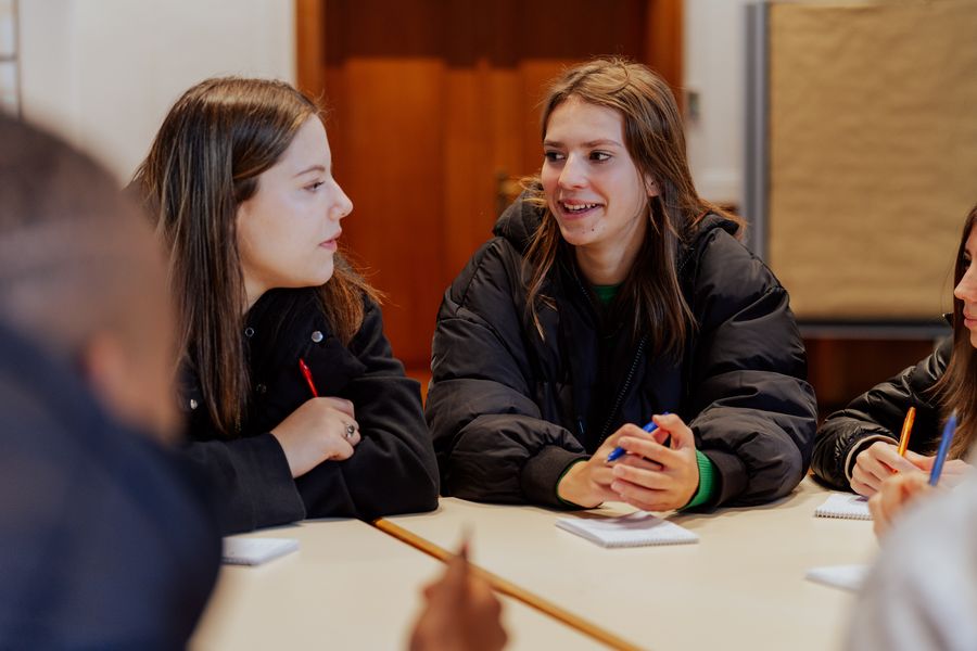 Eine Gruppe von Jugendlichen unterhält sich. Alle haben Kugelschreiber und Papier.