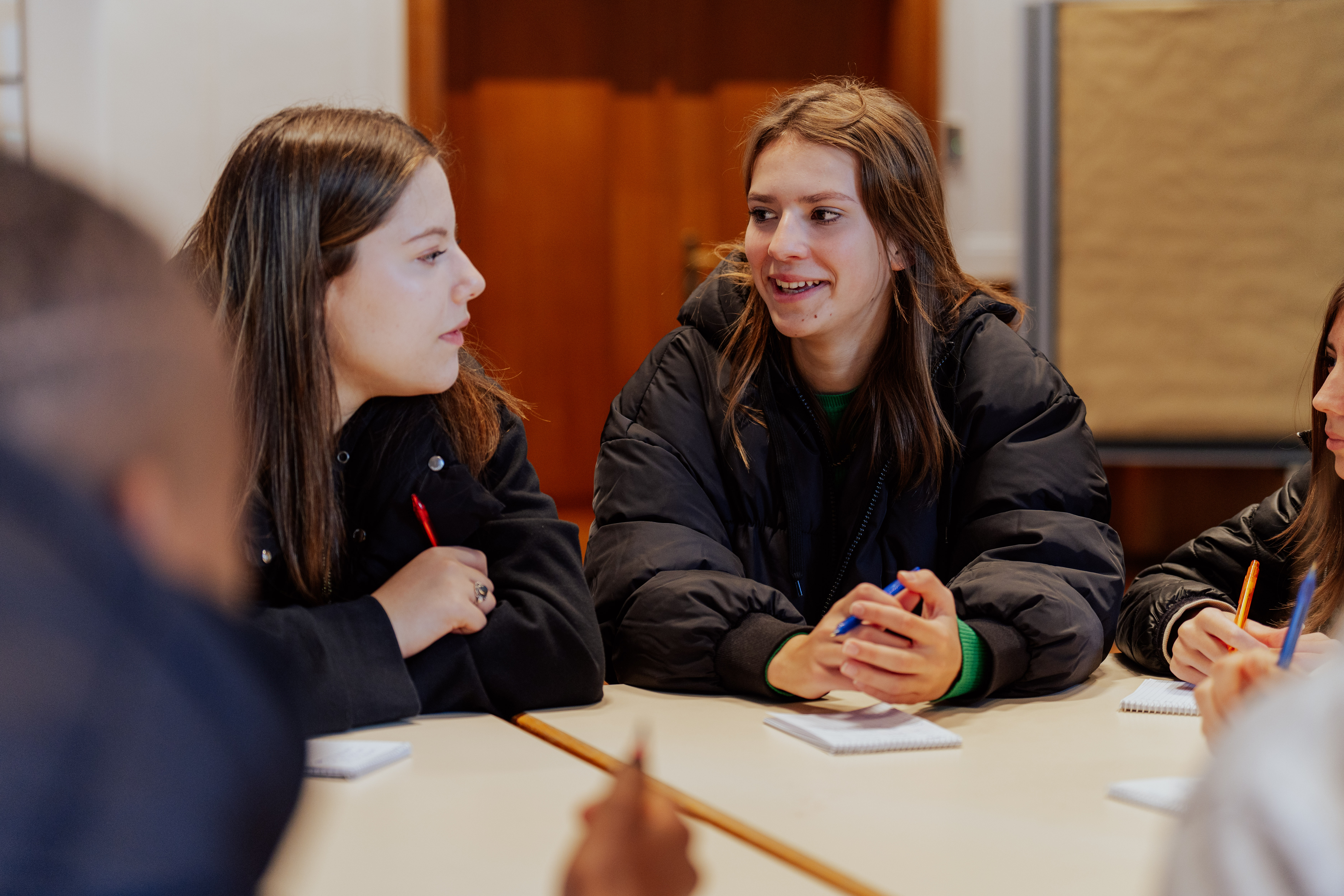 Eine Gruppe von Jugendlichen unterhält sich. Alle haben Kugelschreiber und Papier.