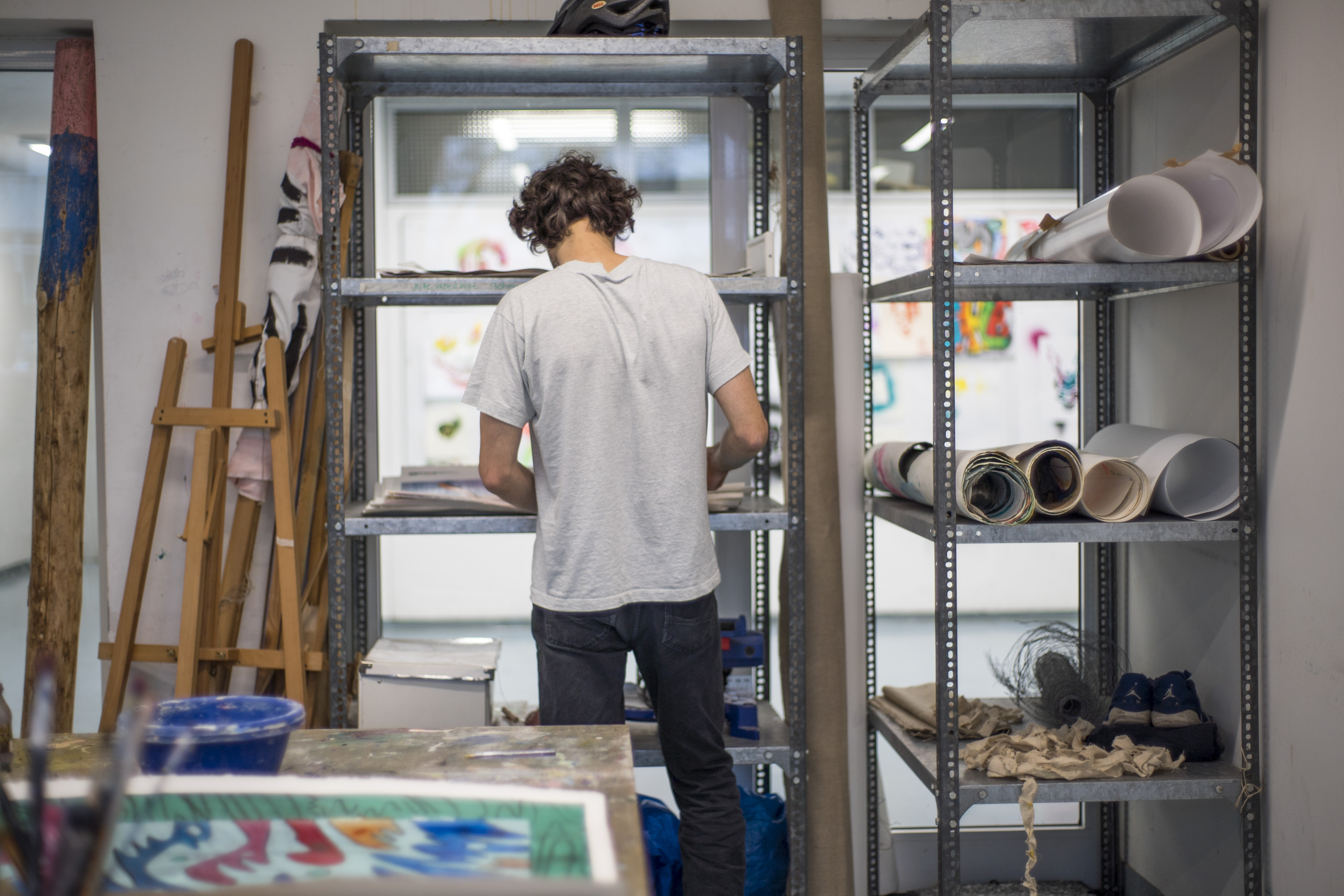 Ein Mann steht vor Regalen mit zusammengerollten Bildern. In der Ecke steht eine Staffelei.